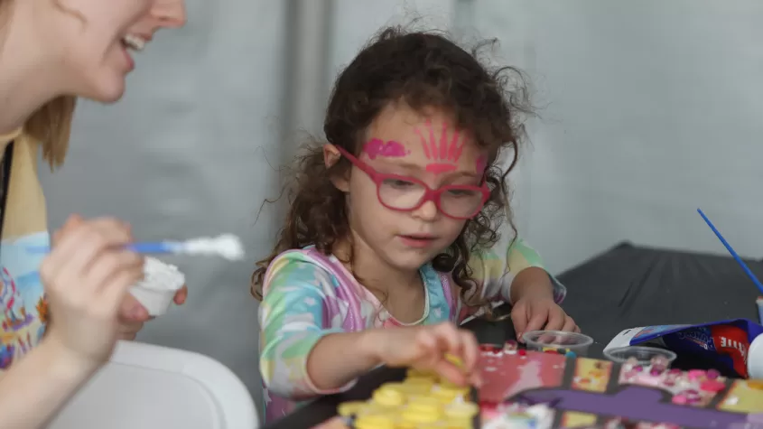 A child participates in a collaborative mosaic activity in our "Kids @ Art" tent, aided by a volunteer.