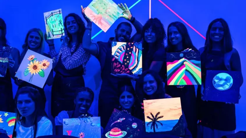 A group of people posing with their glow-in-the-dark paintings under blacklight. The artwork features neon colors, including landscapes, planets, flowers, and abstract designs. The room has a vibrant blue hue with glowing neon lines in the background, creating a fun and creative atmosphere. The participants are smiling and holding up their paintings proudly.