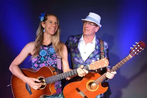 blue background with blonde woman and a man with hat on both smiling while holding a guitar
