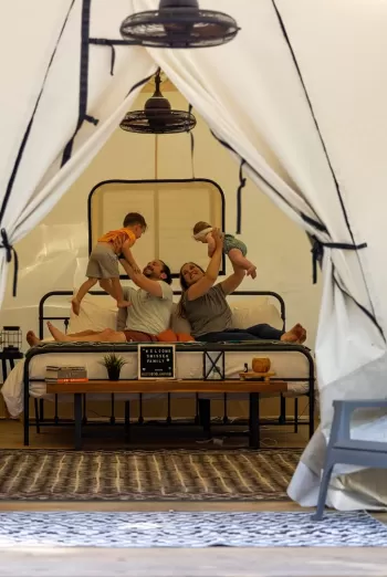 Parents lift their toddler and baby on a bed inside a glamping tent, with a small “Welcome” sign on a bench in front.