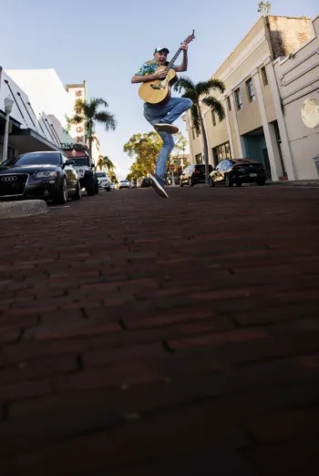 Man jumping with an acoustic guitar on a brick street lined with palm trees and buildings in downtown Fort Myers.