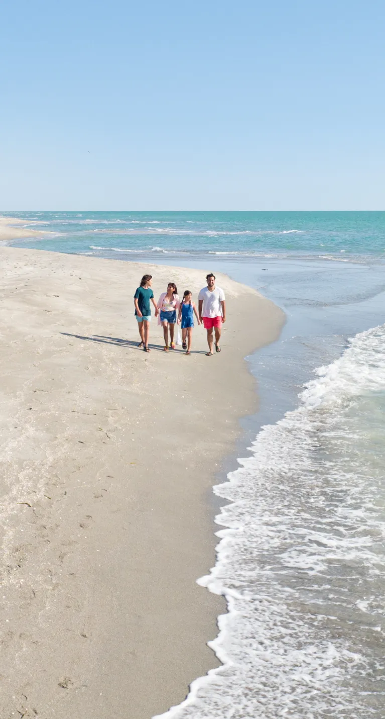 A family of four walking along a wide, empty beach beside calm turquoise water under a clear blue sky.