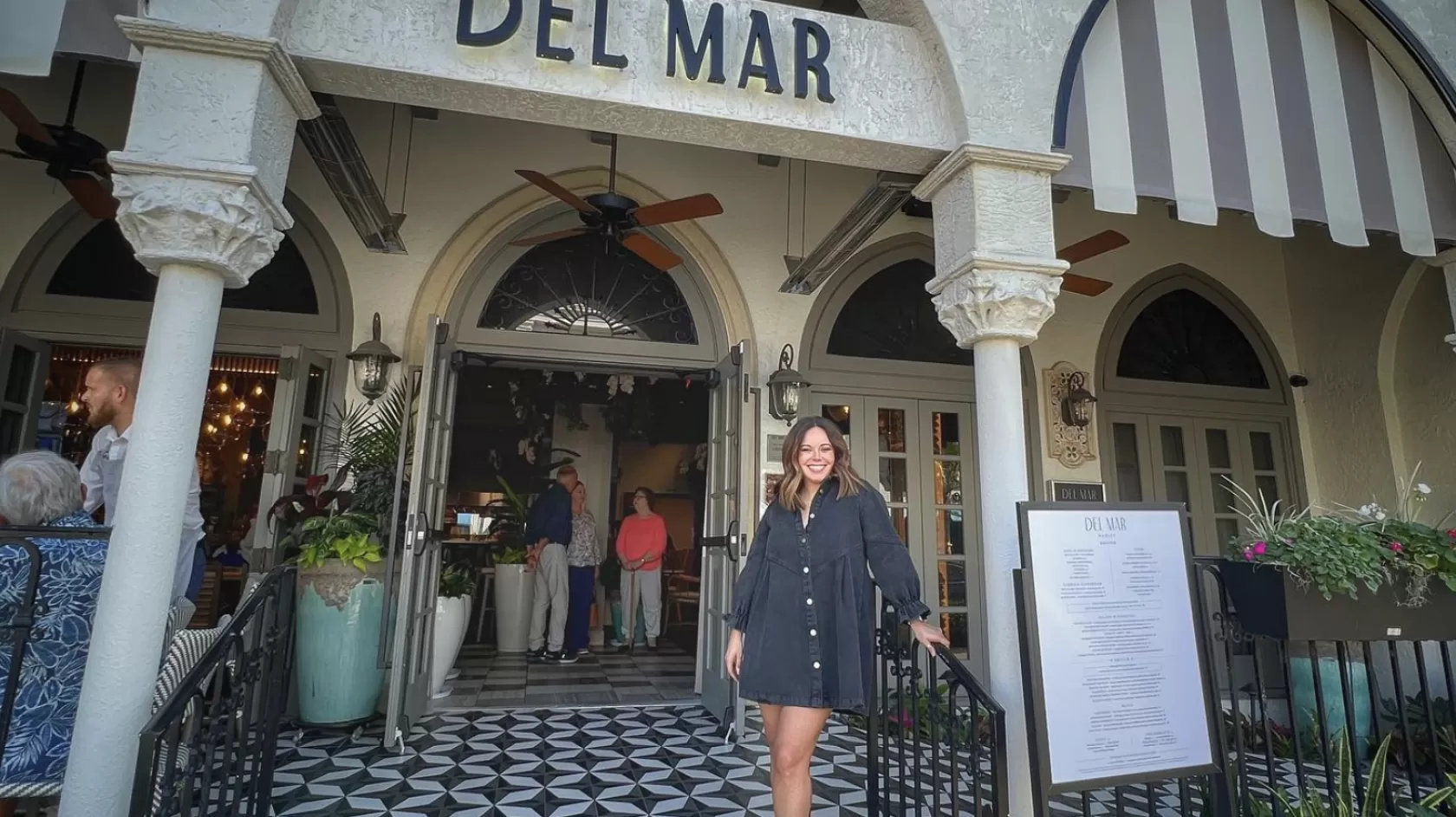A woman stands outside the store Del Mar in a outdoor, mediterranean shopping center. 