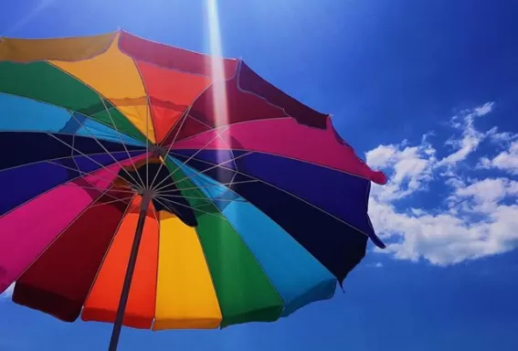 rainbow colored umbrella being viewed frmo underneath with the sun peaking out among a blue sky above it