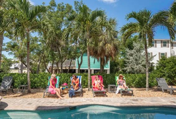 Four women relaxing on lounge chairs beside a pool surrounded by palm trees, chatting and reading in a sunny tropical setting.