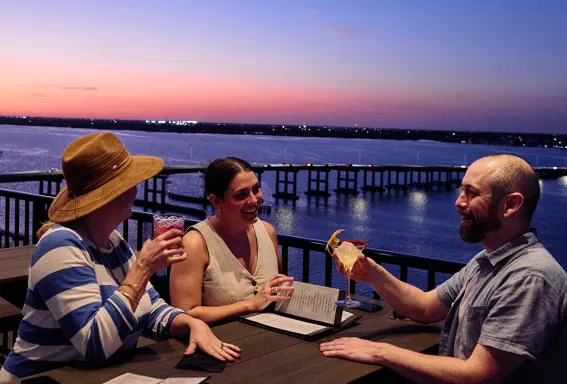 Diners enjoy drinks on a waterfront patio at sunset.