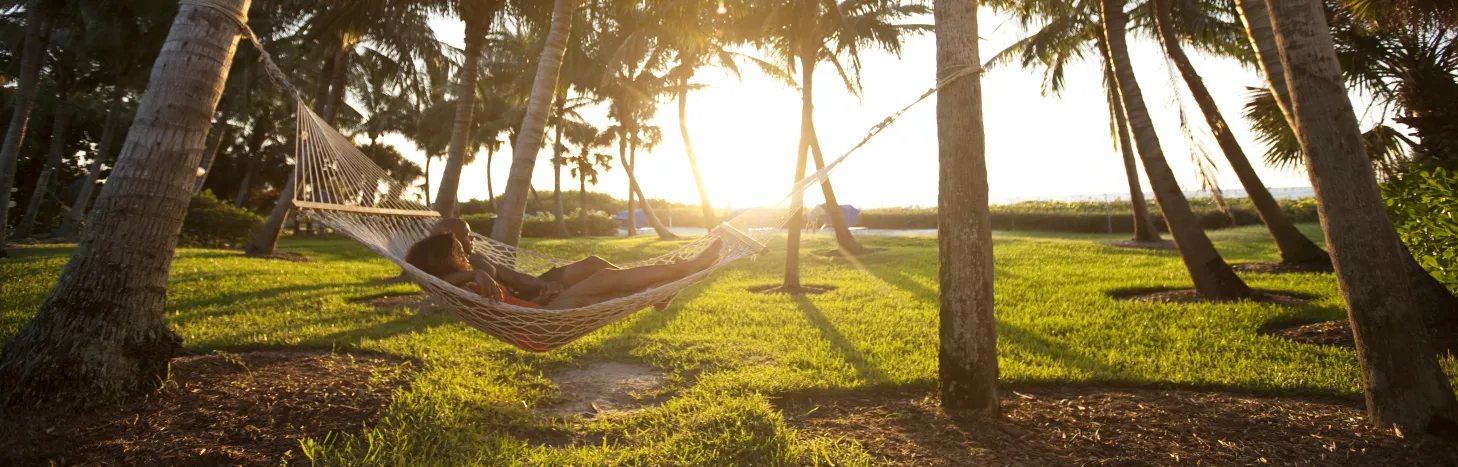 Person relaxing in a hammock between palm trees at sunset with golden light filtering through a tropical lawn near the water.