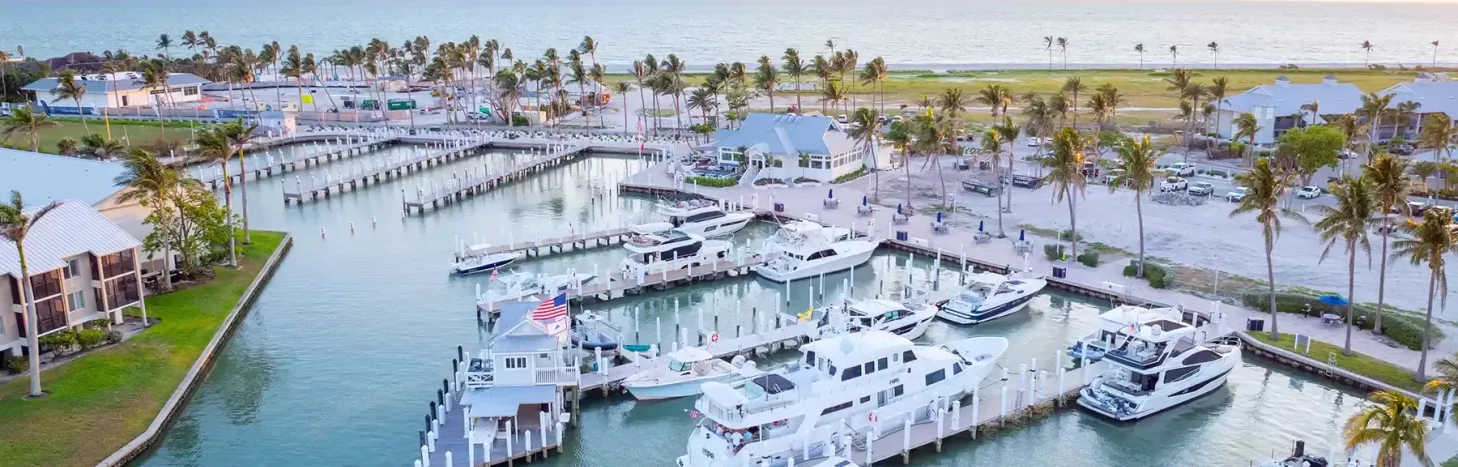 Aerial view of a marina with luxury boats, palm trees, and beachfront buildings along the Gulf Coast.