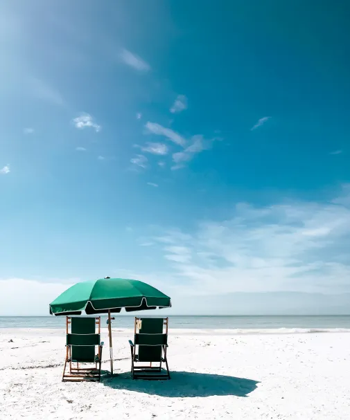 Two green beach chairs under a matching umbrella on a quiet sandy beach facing calm ocean water under a wide blue sky.