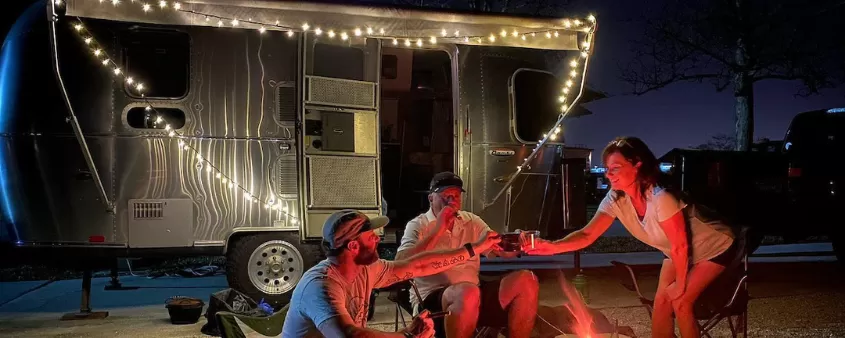 camper with string lights at night; three people around campfire